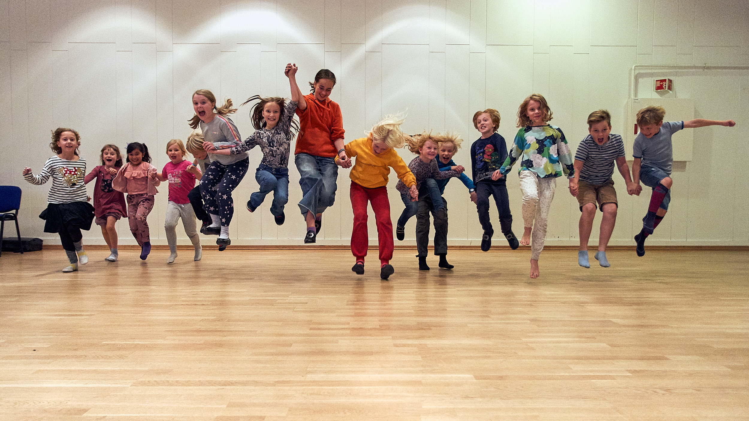 Barn som held hender, står på rekke og hopper.